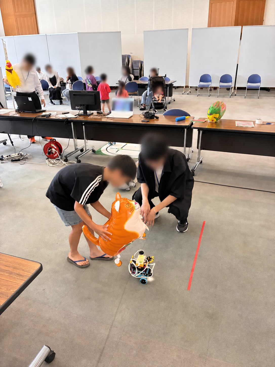 子どもと学生スタッフが、オレンジ色のぬいぐるみと小型ロボットを使っておさんぽを体験している様子